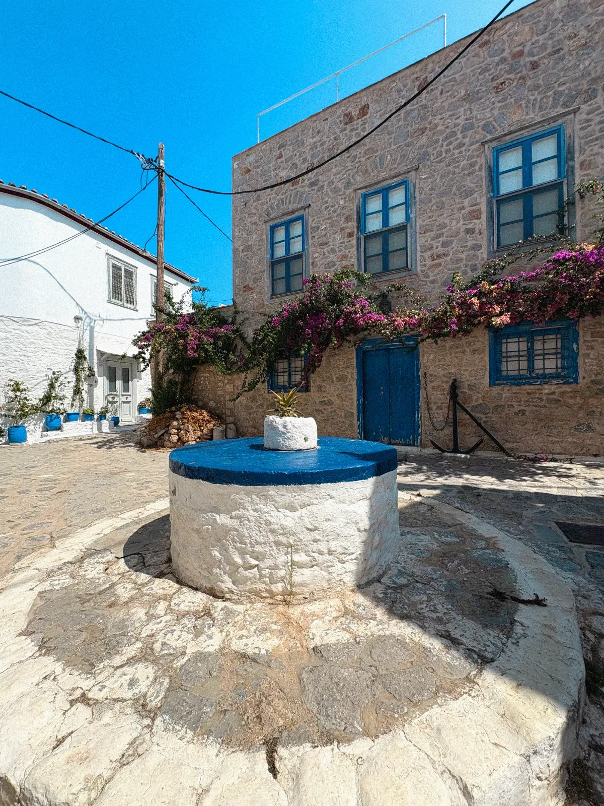 Whitewashed well in a Greek island courtyard beside a stone house with blue shutters